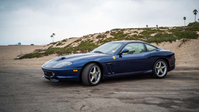 A blue 1999 Ferrari 550 Maranello is parked in front of a sand dune.