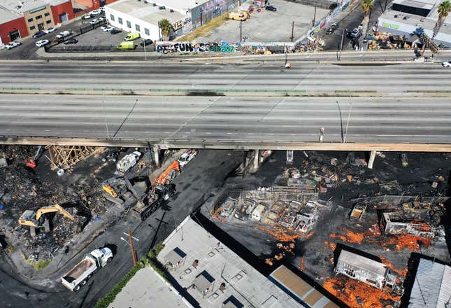 Aerial view of the destruction caused by the pallet fire underneath the 10 freeway in downtown Los Angeles. This image shows the destruction in two storage lots, including lots of charred remains of vehicles and debris including an excavator and the empty freeway.