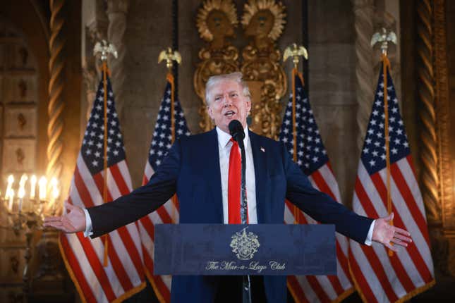 Donald Trump speaking at a podium with his arms out in front of four American flags