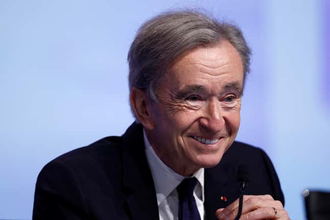 Bernard Arnault in a suit, at a microphone, in front of a screen, smiling