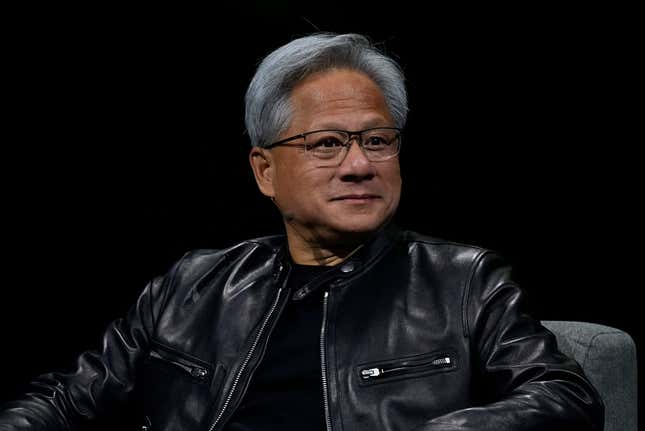 Jensen Huang smiling while wearing a black leather jacket sitting in a grey chair in front of a black backdrop