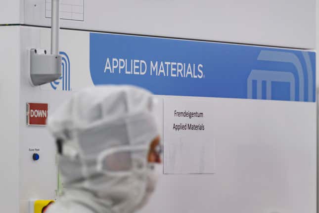 Una persona que lleva un casco protector y una máscara camina en primer plano mientras que en el fondo hay un cartel de Applied Materials en la pared.