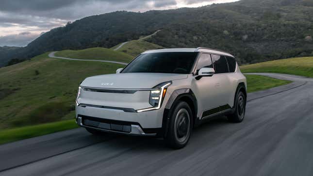 A Kia EV9 driving down a two-line road across hilly terrain.