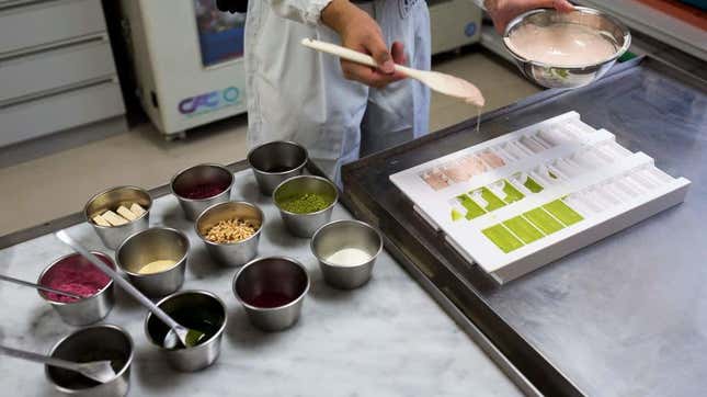 Green and peach-colored Kit Kat chocolate being poured into candy molds beside metal canisters full of ingredients