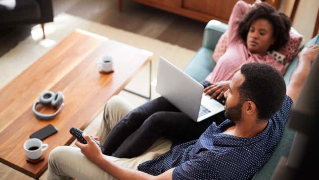 Couple Sitting On Sofa At Home Using Laptop Computer And Watching TV 